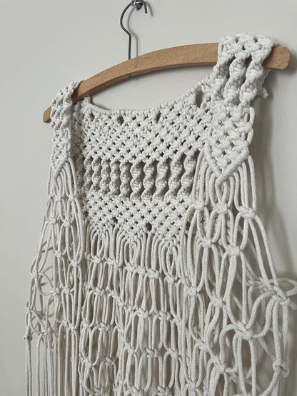 Close up of the intricate knot detail on a handmade natural boho fringed waistcoat hanging on a wooden coat hanger against a beige wall. 