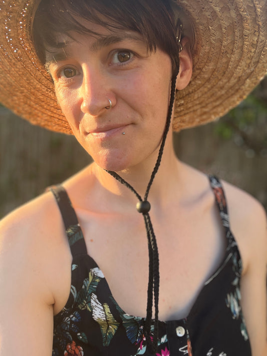 A woman with short brown hair wearing a straw hat and floral dress with a blurred natural background, is looking towards the camera. She has a black chin strap attached to her hat, which is secured under the chin with a black toggle. 
