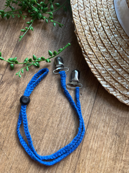Blue braided macrame chin strap with metal clips and a black toggle on a wooden surface next to a straw hat and plant.