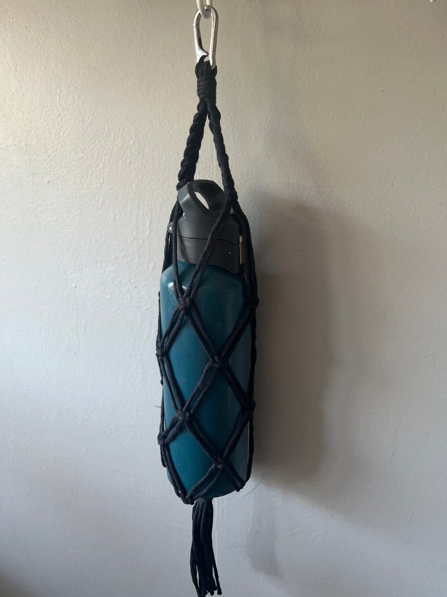 Black macrame bottle holder, holding a blue water bottle securely inside, with a metal carabiner clip at the top and a tassel at the bottom. 
