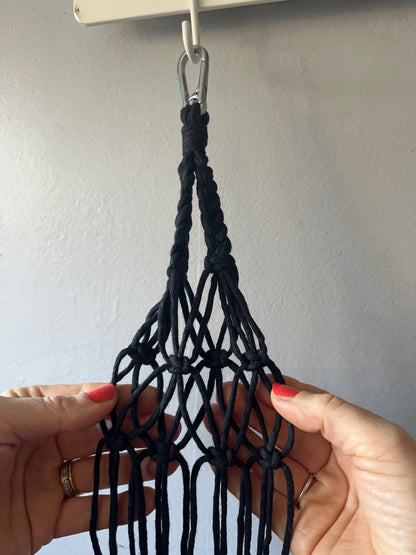 Hands holding a partially completed project made with black macrame yarn on a silver carabiner clip, against a grey background. 