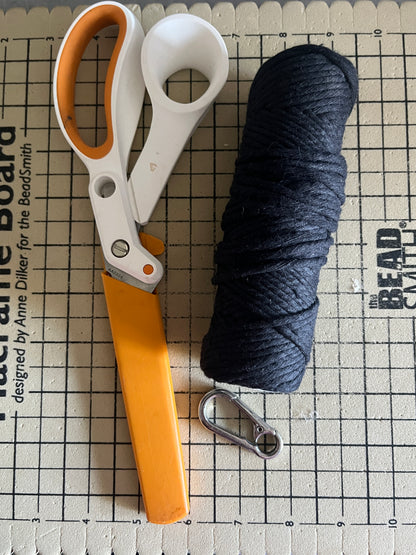 Roll of black macrame yarn, scissors with orange handles, and a metal carabiner, laid out on a grid surface.