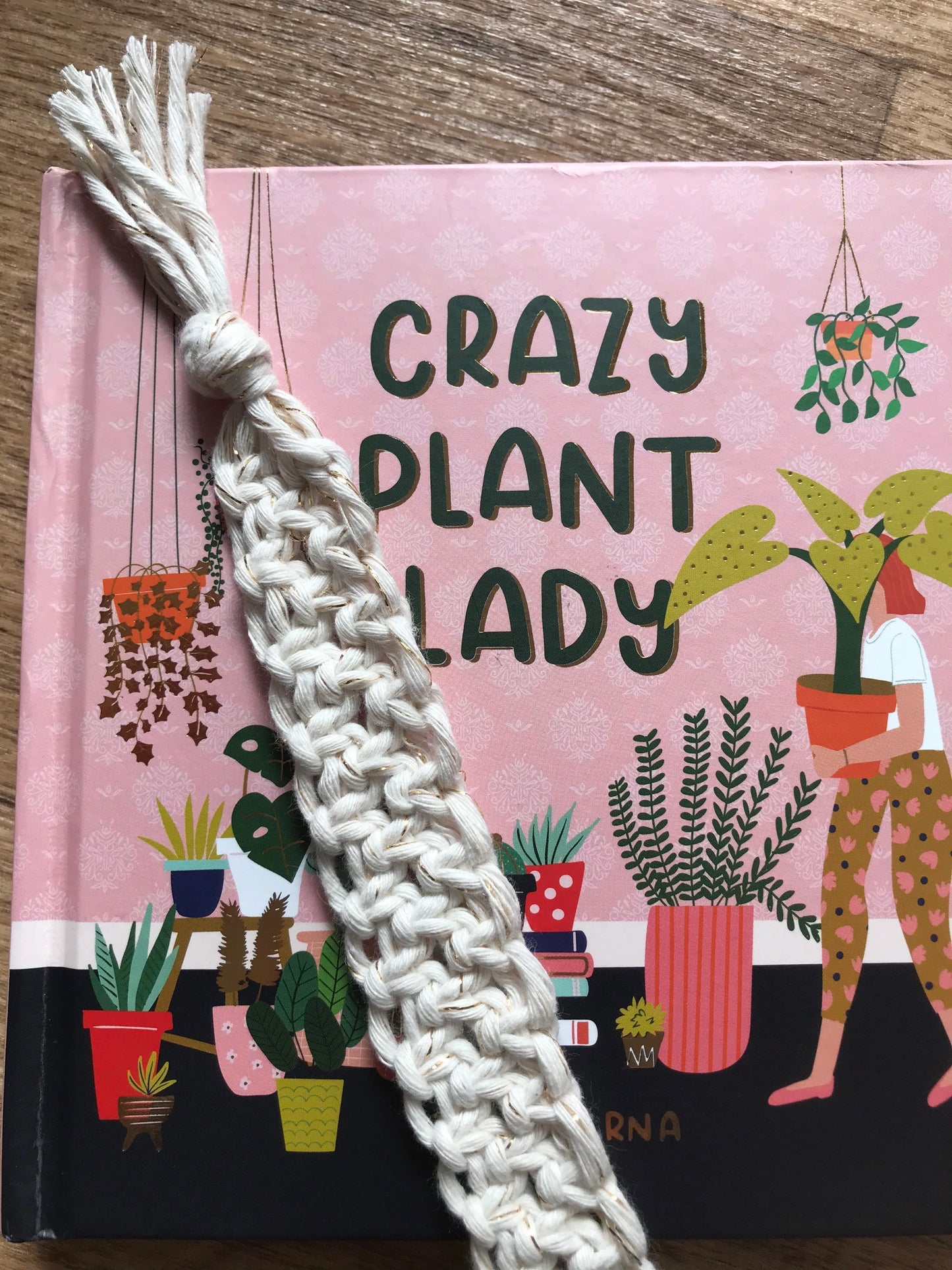 Handmade macramé bookmark with knotted design made with golden natural recycled cotton, laid across a closed hardback book entitled "Crazy Plant Lady" on a wooden surface.