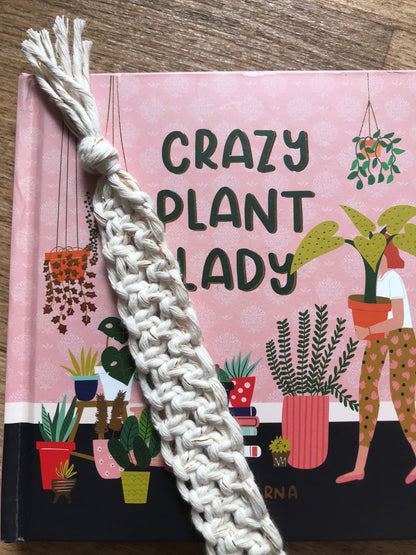Handmade macramé bookmark with knotted design made with golden natural recycled cotton, laid across a closed hardback book entitled "Crazy Plant Lady" on a wooden surface.