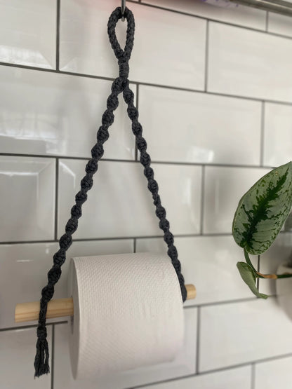 A roll of toilet paper hangs on a wooden dowel, suspended by a Macra-Made-With-Love Single toilet roll holder. This bathroom accessory, attached to a white tiled wall, adds an eco-friendly decor touch with a green potted plant visible to the right.