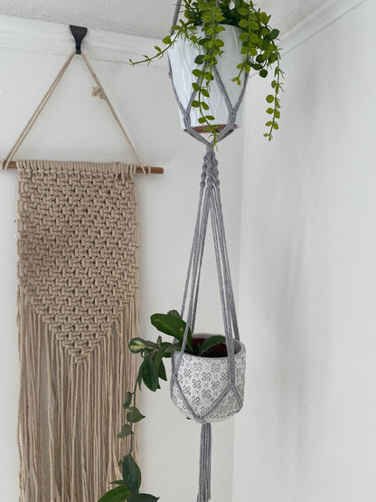 Close up of two green plants in ceramic pots, suspended vertically in a large double macramé plant hanger, made with light grey yarn in the corner of a room with white walls. There is a macrame wall hanging in the background. 
