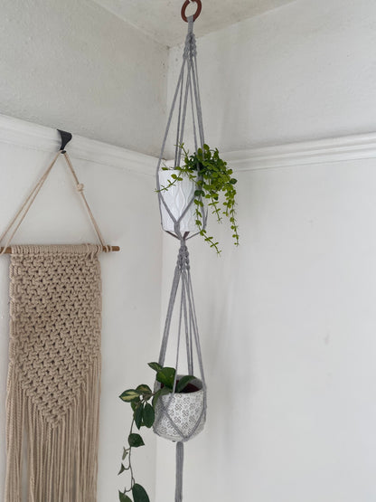 Two green plants in ceramic pots, suspended vertically in a large double macramé plant hanger, made with light grey yarn and a wooden hanging ring, in the corner of a room with white walls. There is a macrame wall hanging in the background. 