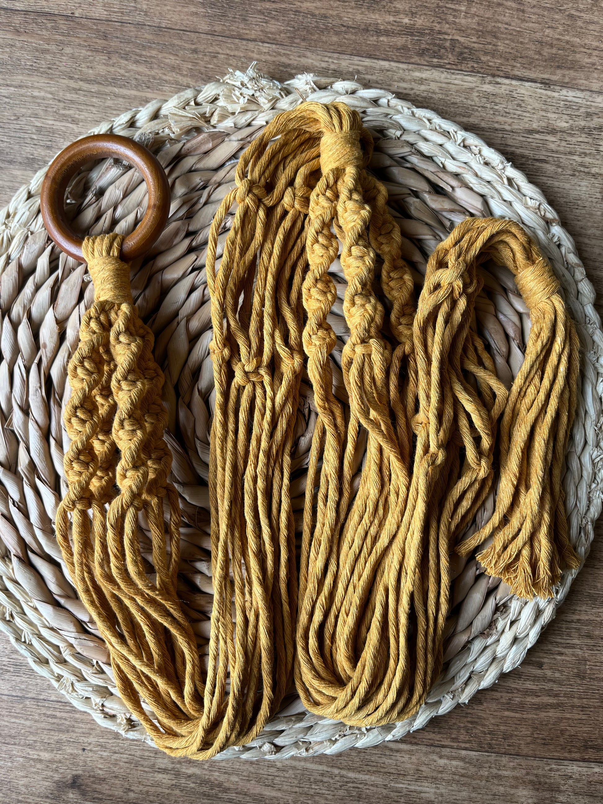A handmade macrame plant hanger made with mustard yellow yarn and a wooden ring, is folded neatly on a round wicker placemat. 