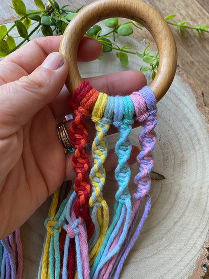 A hand holding a wooden ring at the top of a handmade macrame plant hanger, made with rainbow coloured yarn. 