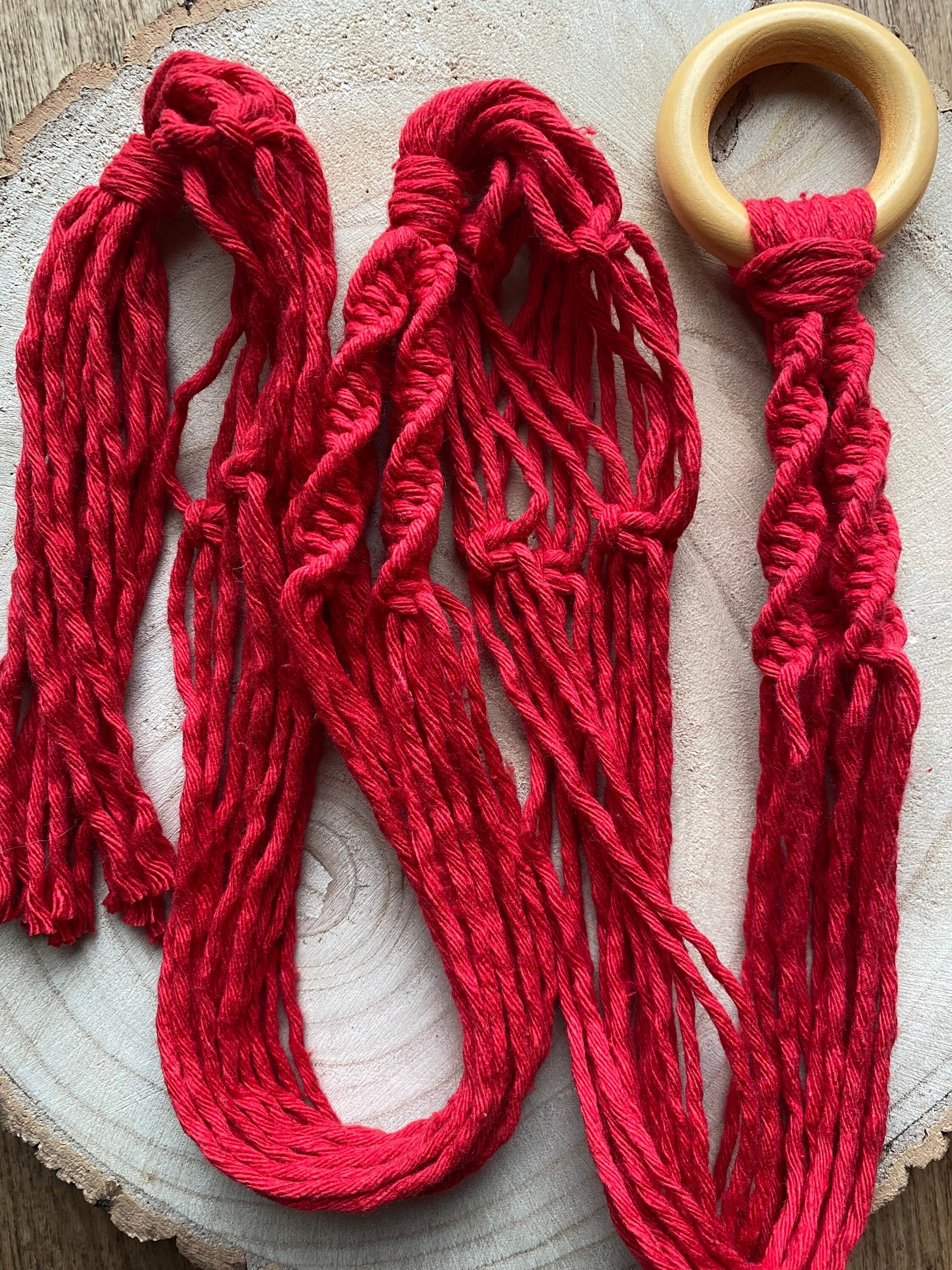A macrame plant hanger made with red yarn and a wooden ring, lays coiled on a wooden surface. 