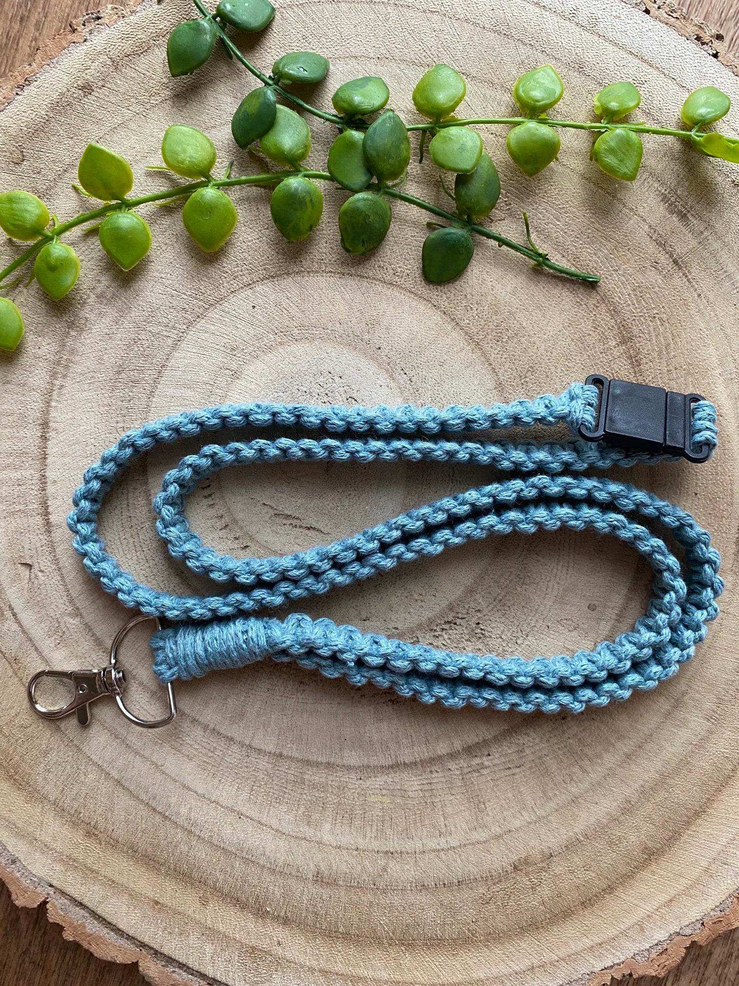 A handmade light blue Eco friendly lanyard badge holder - 34” by Macra-Made-With-Love with a silver clip and a black plastic clasp is coiled neatly on a wooden surface. Above the eco-friendly lanyard, a small branch with green leaves lies decoratively on the wood.