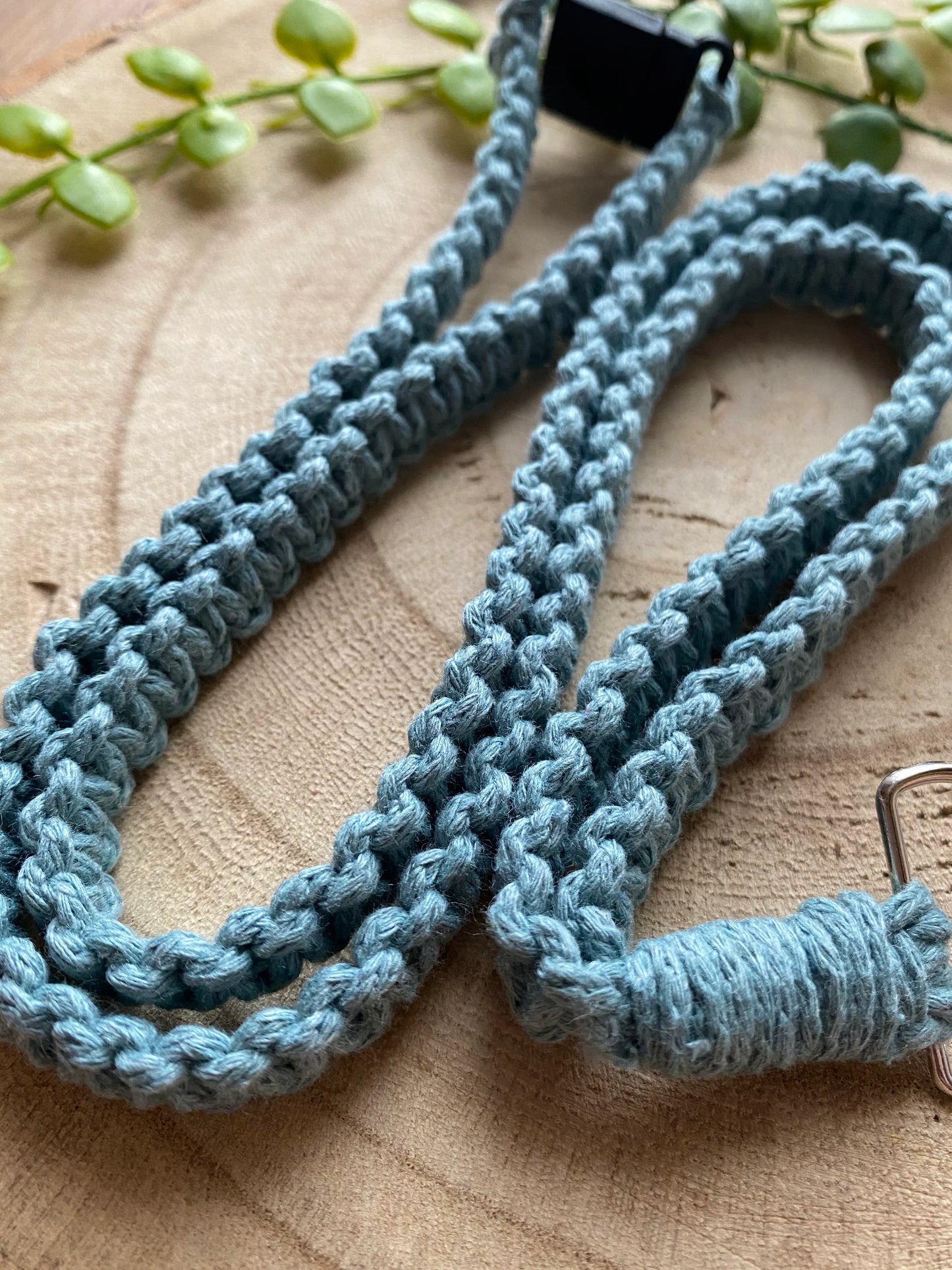 Close-up of an Eco friendly lanyard badge holder - 34”, crafted from blue braided material, placed on a wooden surface with a green plant in the background. One end features a black clip, and the other end has a metal ring. Brand name: Macra-Made-With-Love.