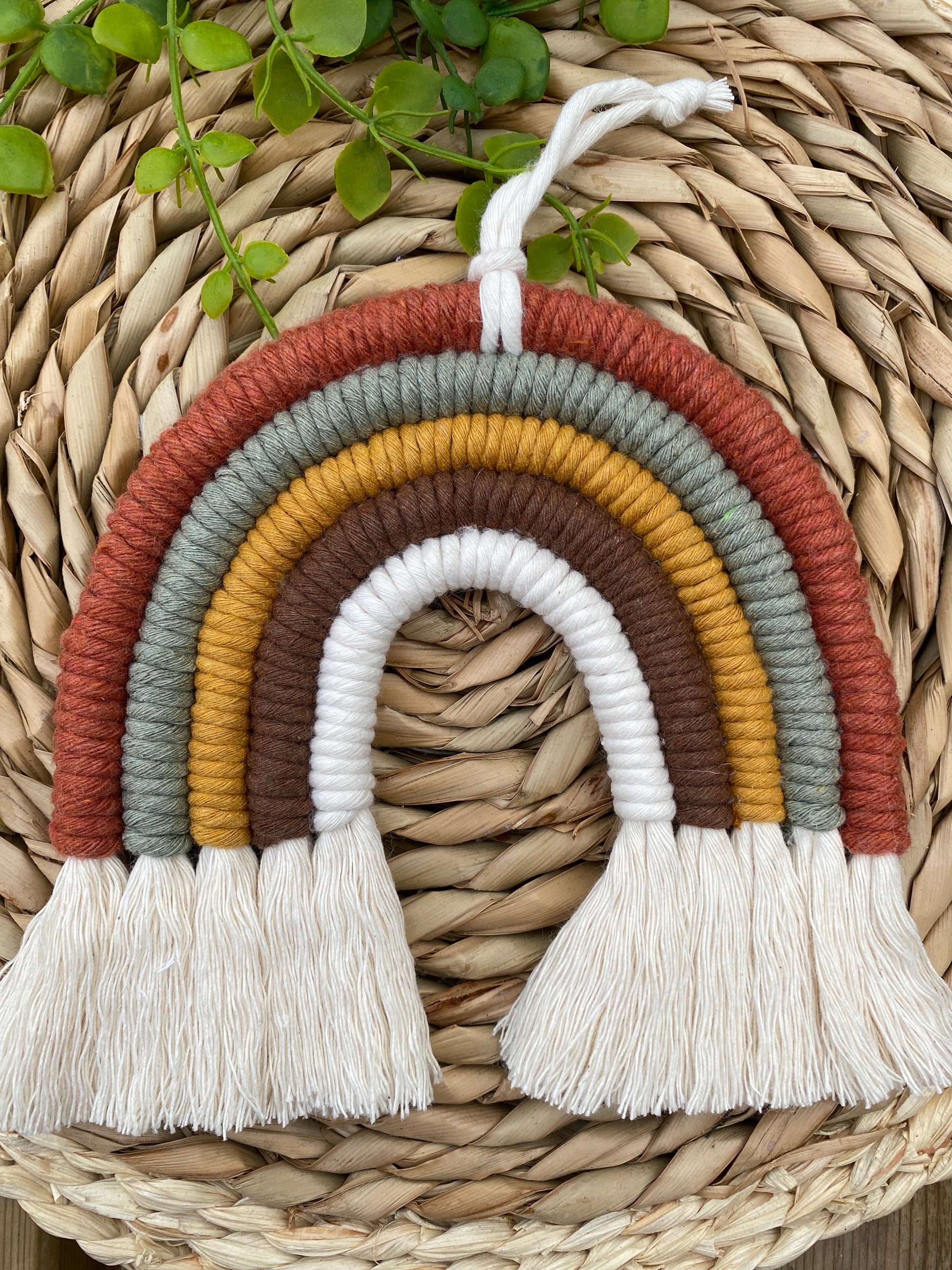 A handmade macrame rainbow, in earthy brown, mustard, green tones, with natural fringed ends, placed on a wicker placemat with some green plant vines. 