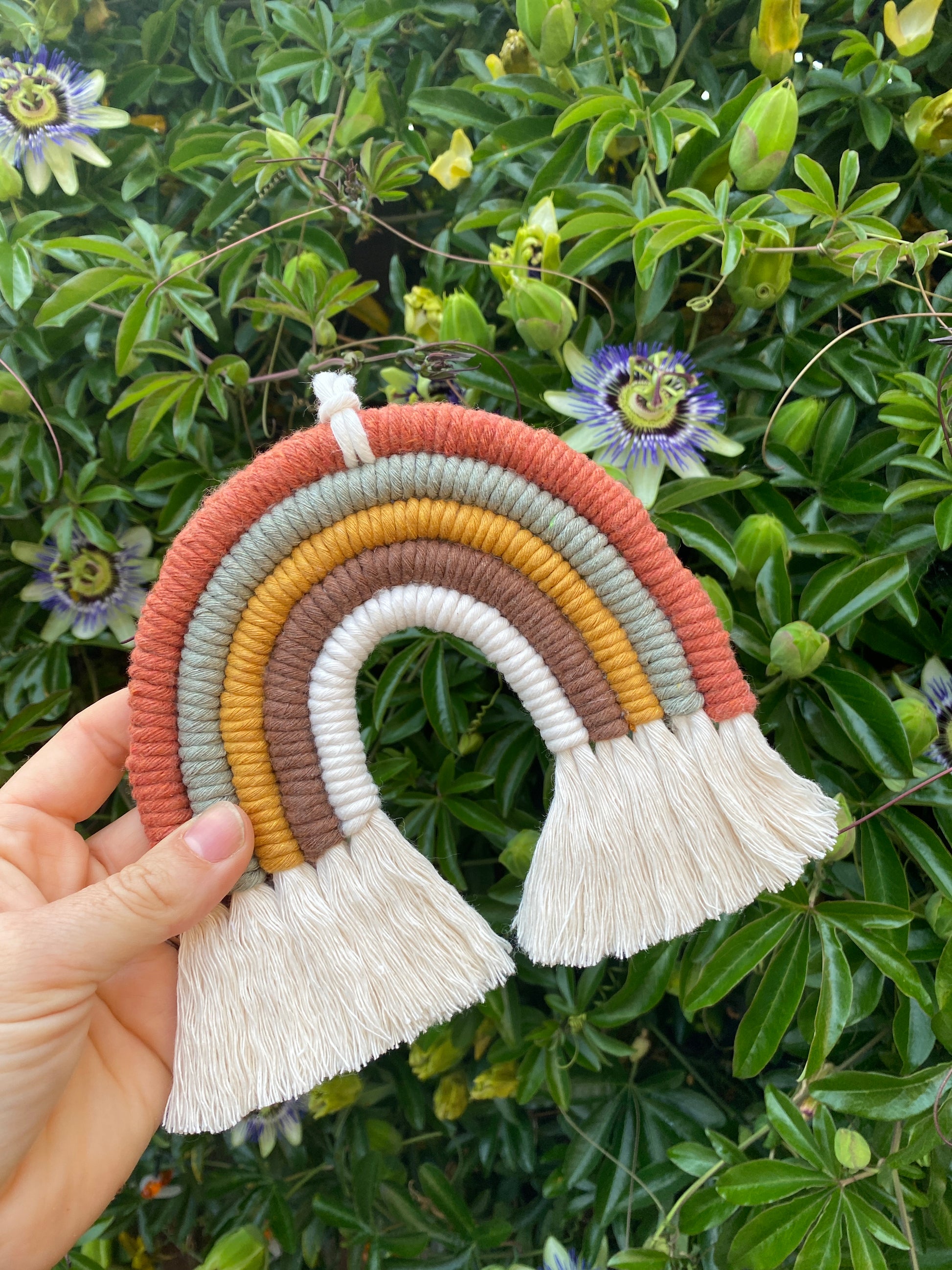 A hand holding a handmade macrame rainbow, in earthy brown, mustard, green tones, with natural fringed ends, outside with leafy green plants in the background. 