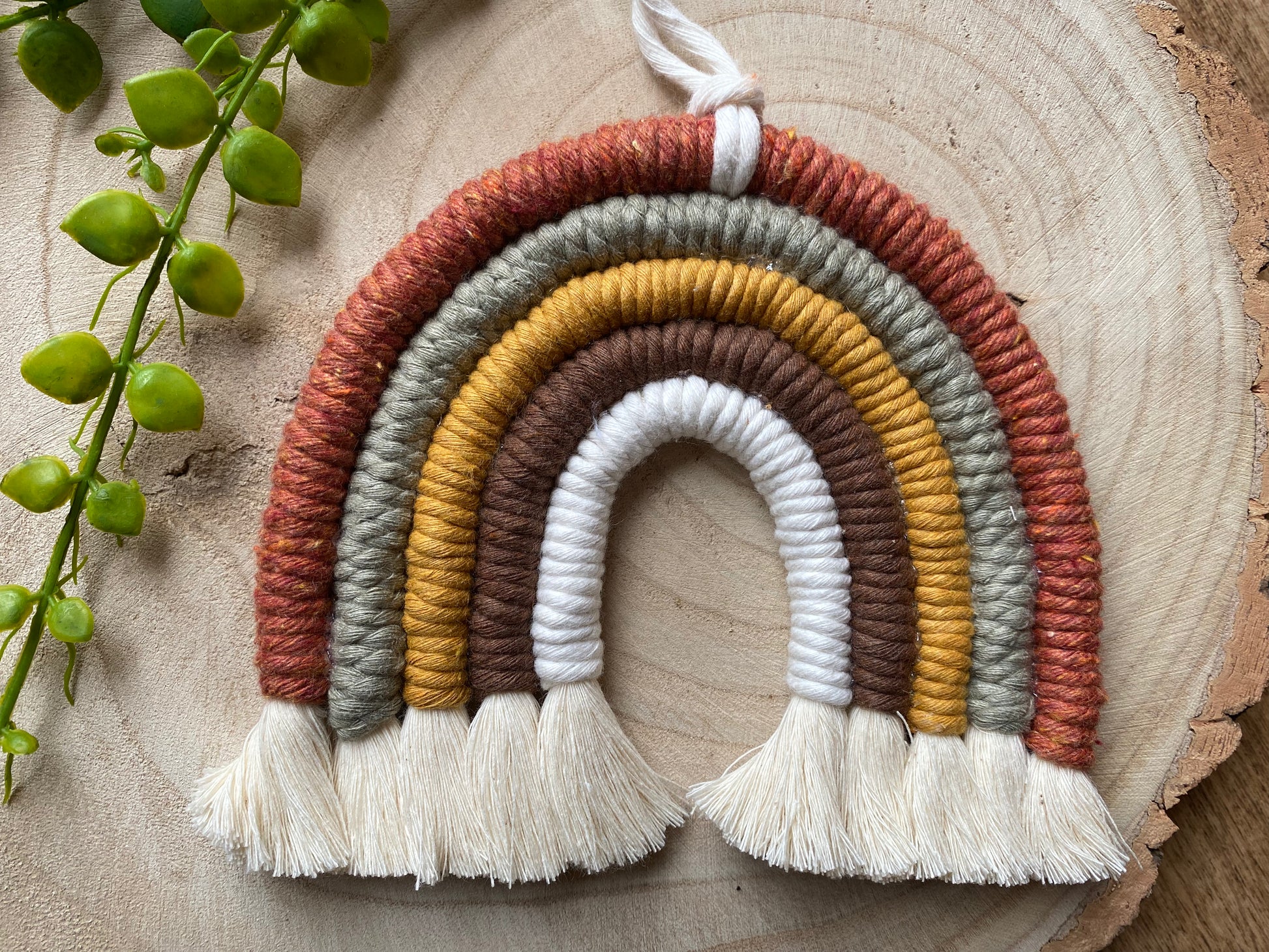 A handmade macrame rainbow, in earthy brown, mustard, green tones, with natural fringed ends, placed on a wooden placemat with some green plant vines. 