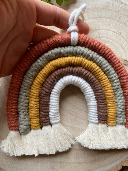 A hand holding a handmade macrame rainbow, in earthy brown, mustard, green tones, with natural fringed ends.