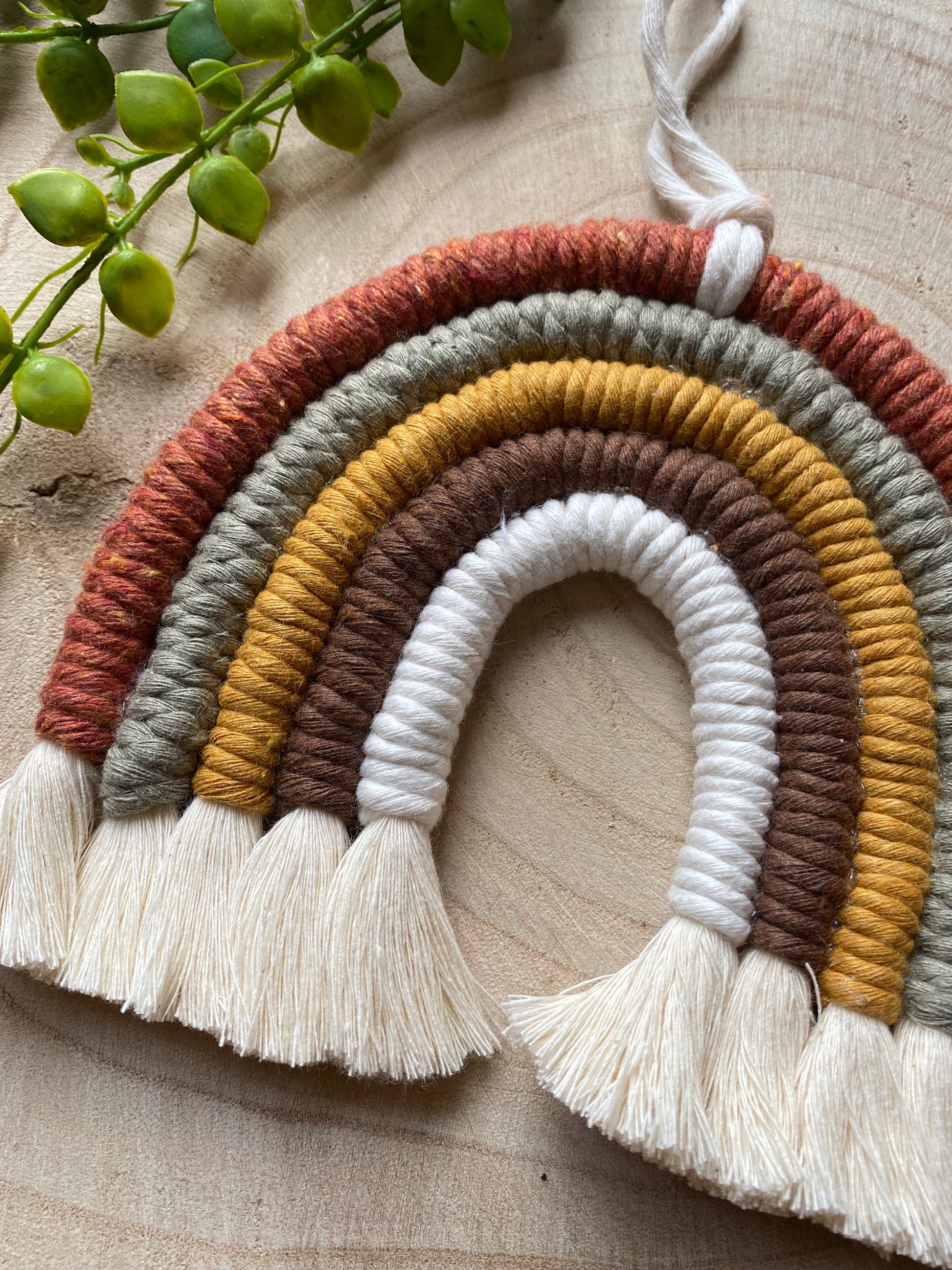 A close up of a handmade macrame rainbow, in earthy brown, mustard, green tones, with natural fringed ends, placed on a wooden placemat with some green plant vines. 