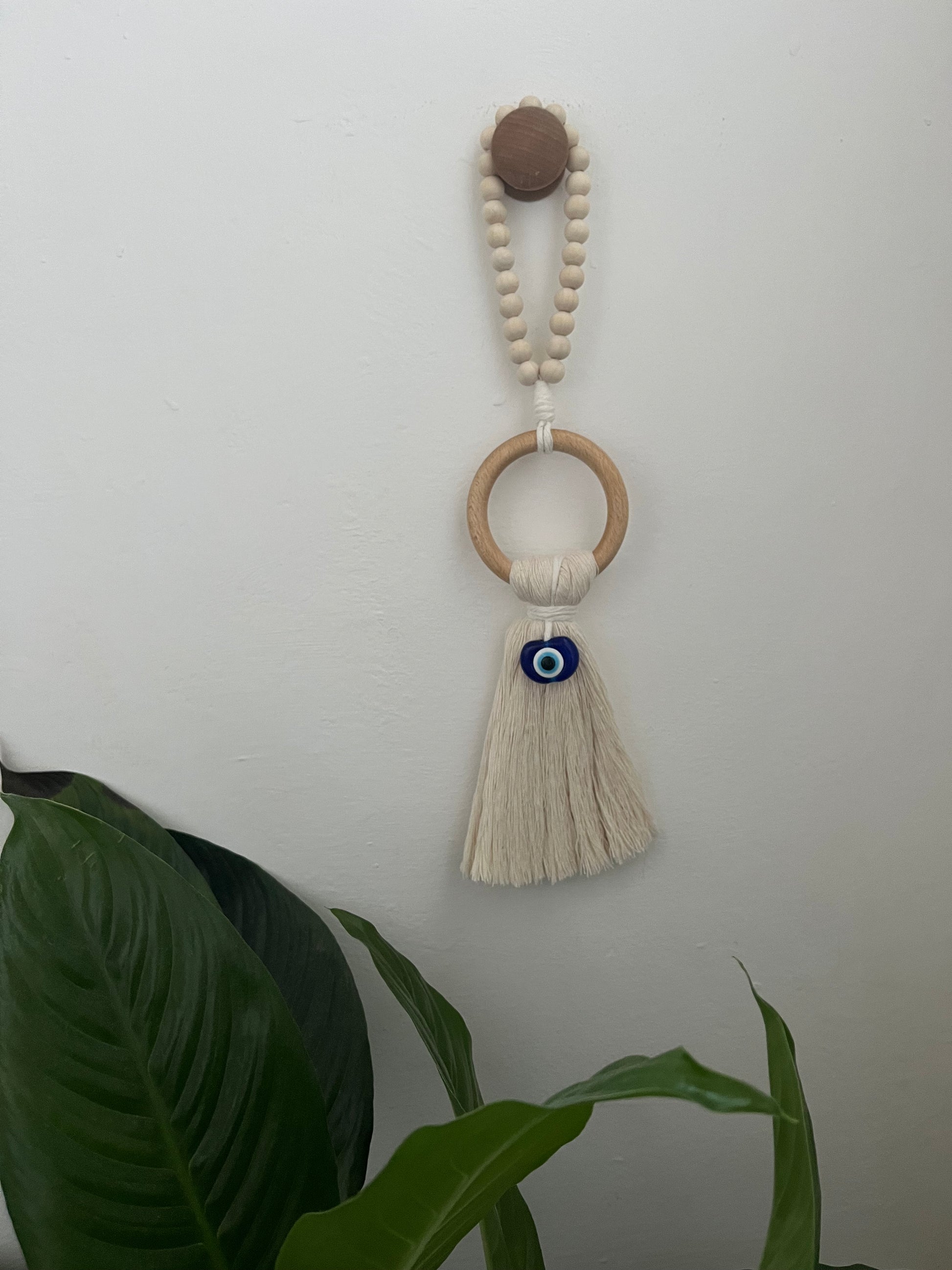 Handmade macrame door hanger, made with recycled cotton, on a wooden ring, with a blue resin evil eye bead and beaded hanging loop. Shown hanging from a wooden knob on a white wall. 