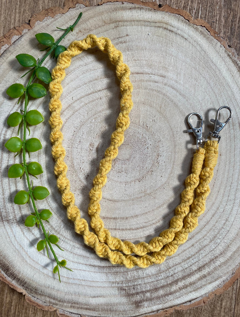 Handmade macramé face mask lanyard in mustard yellow cotton cord with small metal clips on each end, on a round wooden surface. 