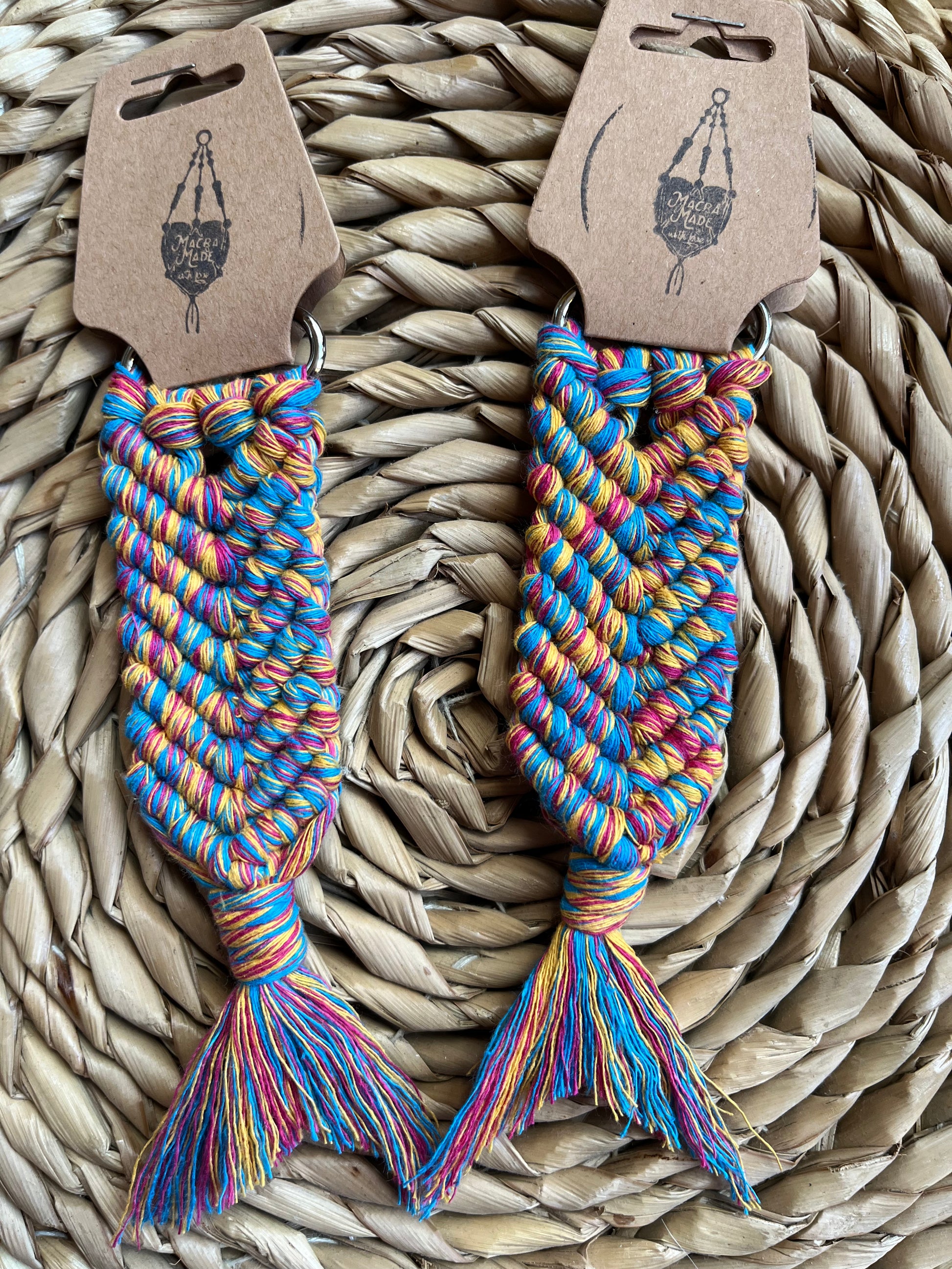 Two handmade macrame keyrings, knotted using brightly coloured recycled cotton, in the shape of a mermaids tail. Each keyring has a brown card sing tag attached, with the Macra-made With Logo stamped on the tag. 