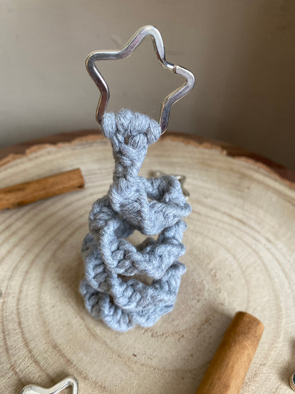 Close up of a freestanding spiral Christmas tree ornament made with macrame knots in grey yarn with a silver star on top, displayed on a wooden surface with cinnamon sticks for decoration. 