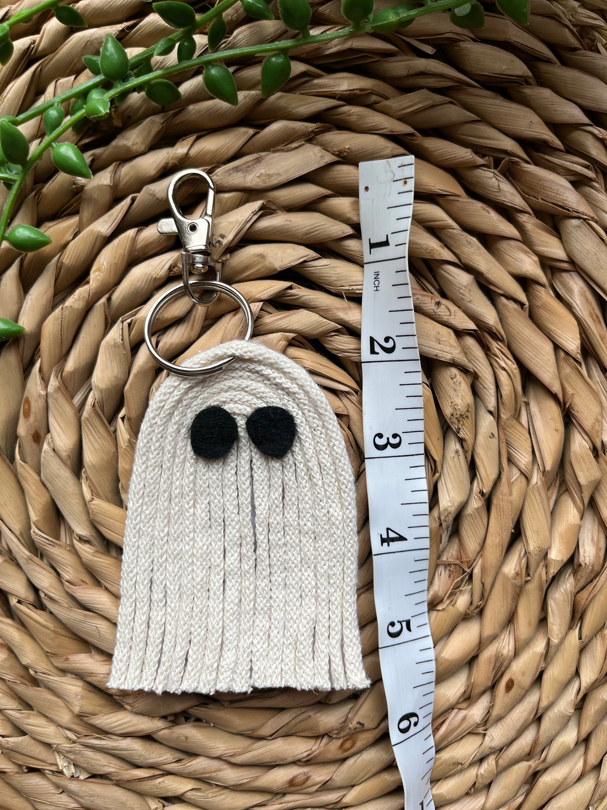 White ghost shaped keychain with black eyes and a silver keyring clasp, laid out on a woven mat next to a white tape measure. 
