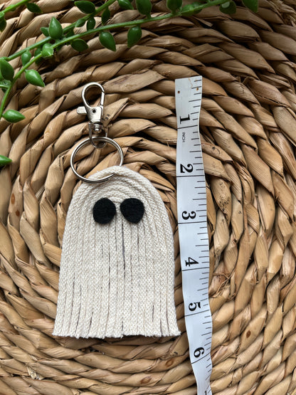 White ghost shaped keychain with black eyes and a silver keyring clasp, laid out on a woven mat next to a white tape measure. 