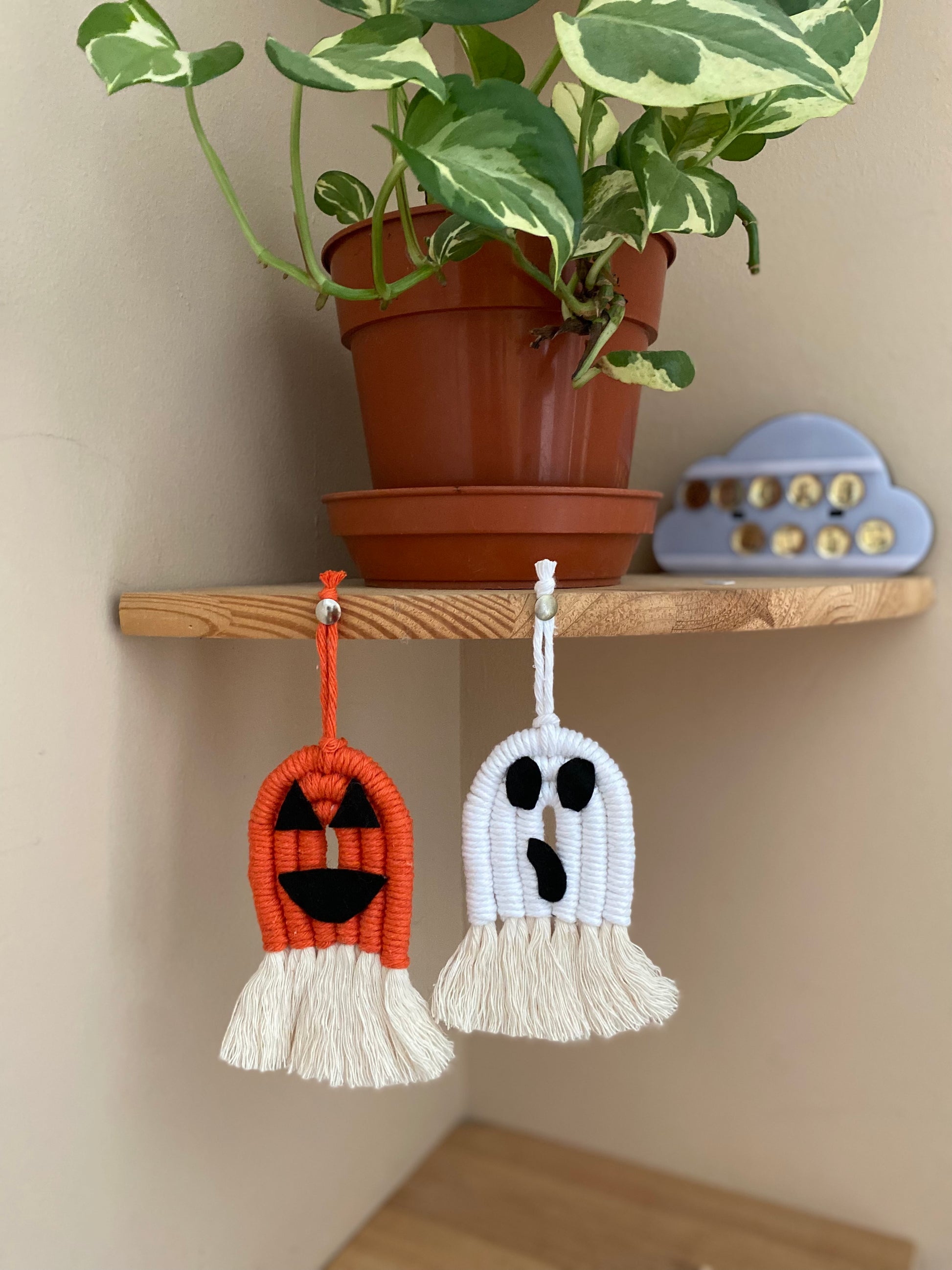 A pair of macrame ghost and pumpkin ornaments, made with orange and white yarns with black felt face details and long natural tassels at the bottom, hanging on the edge of a wooden corner shelf, with a variegated plant in a plastic pot on the shelf above. 