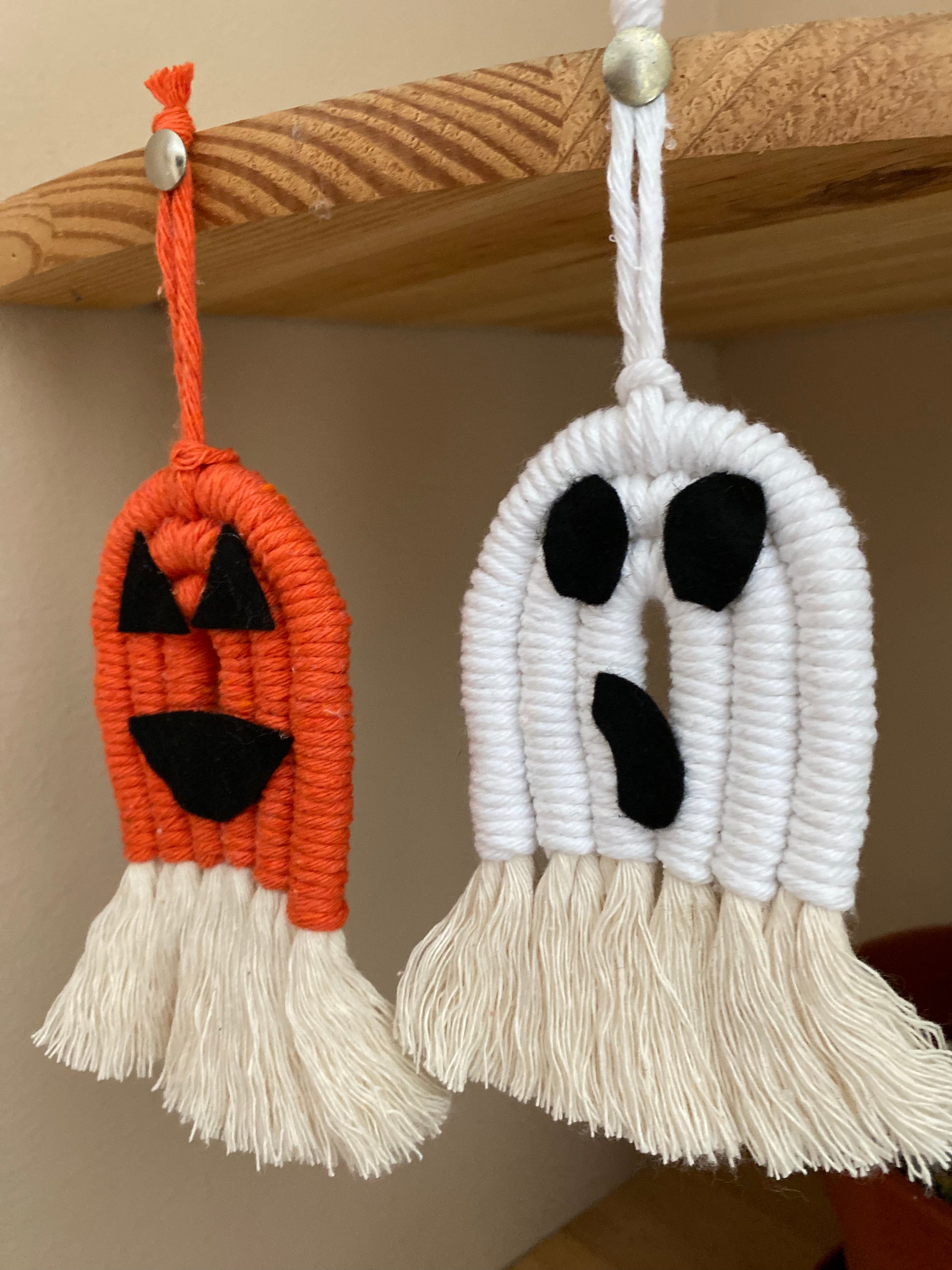 A set of macrame ghost and pumpkin ornaments, made with orange and white yarns with black felt face details and long natural tassels at the bottom, hanging with pins on the edge of a wooden shelf. 