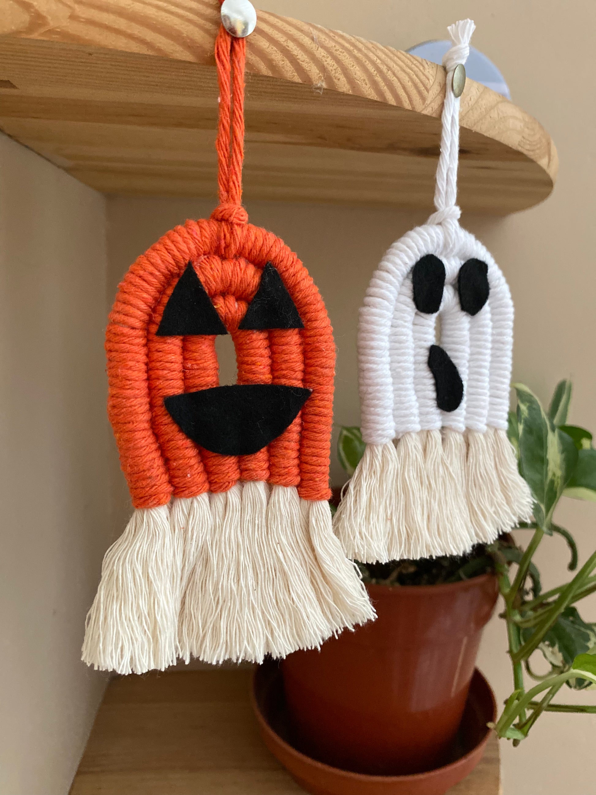 A pair of macrame ghost and pumpkin ornaments, made with orange and white yarns with black felt face details and long natural tassels at the bottom hanging in front of a houseplant from the edge of a wooden shelf. 