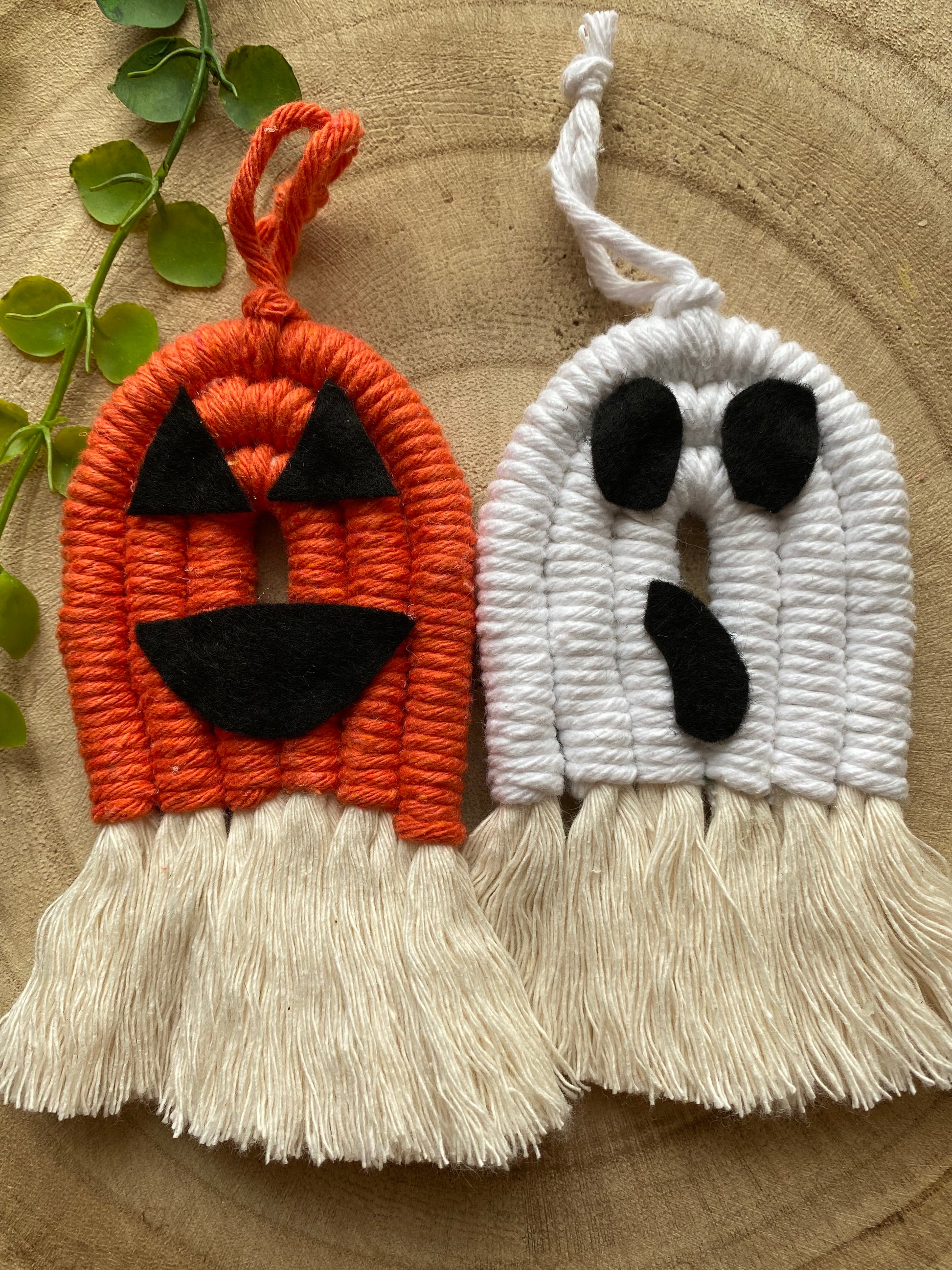 A pair of macrame ghost and pumpkin ornaments, made with orange and white yarns with black felt face details and long natural tassels at the bottom, displayed next to each other on a wooden board. 