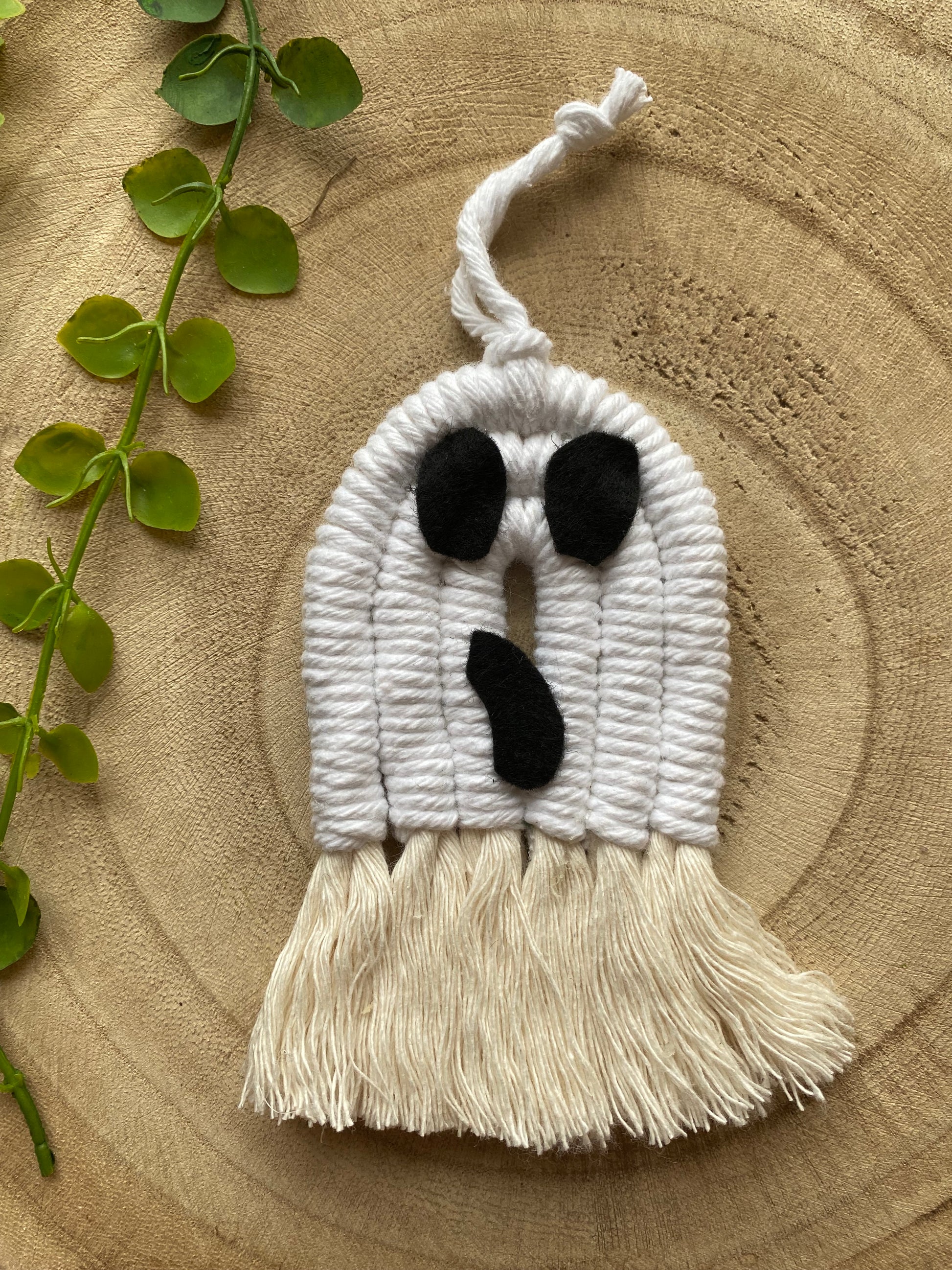 A macrame ghost ornament, made with white yarn with black felt face details and long natural tassels at the bottom, on a wooden board. 