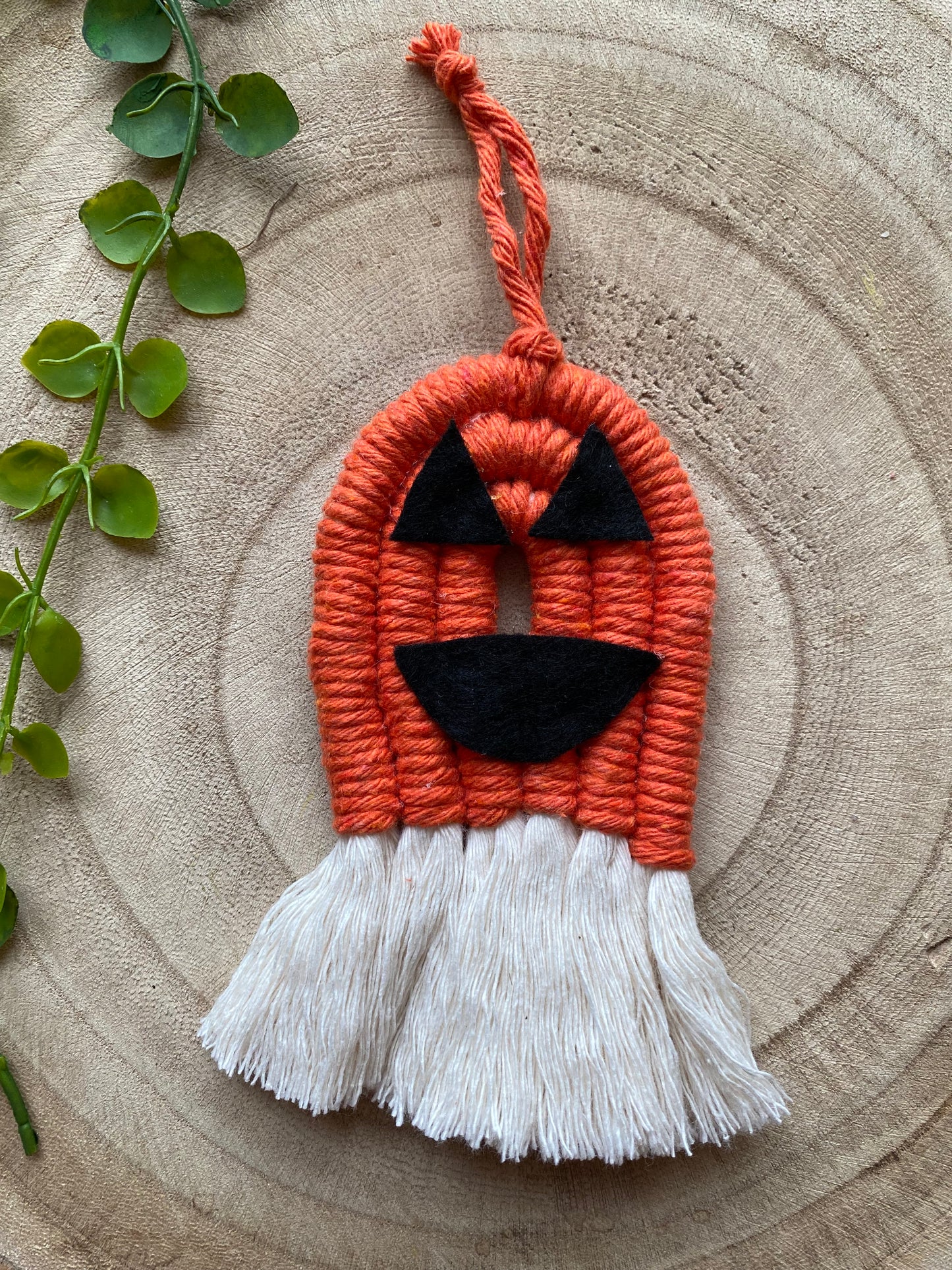A macrame pumpkin ornament, made with orange yarn with black felt face details and long natural tassels at the bottom on a wooden board. 