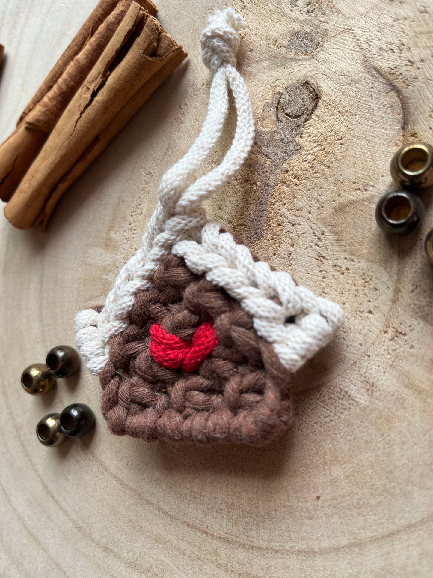 Close up side angle of a small macramé decoration in the shape of a gingerbread house, made with brown and white yarn with a red heart detail in the middle, styled on a light wood surface with cinnamon sticks and metal beads. 