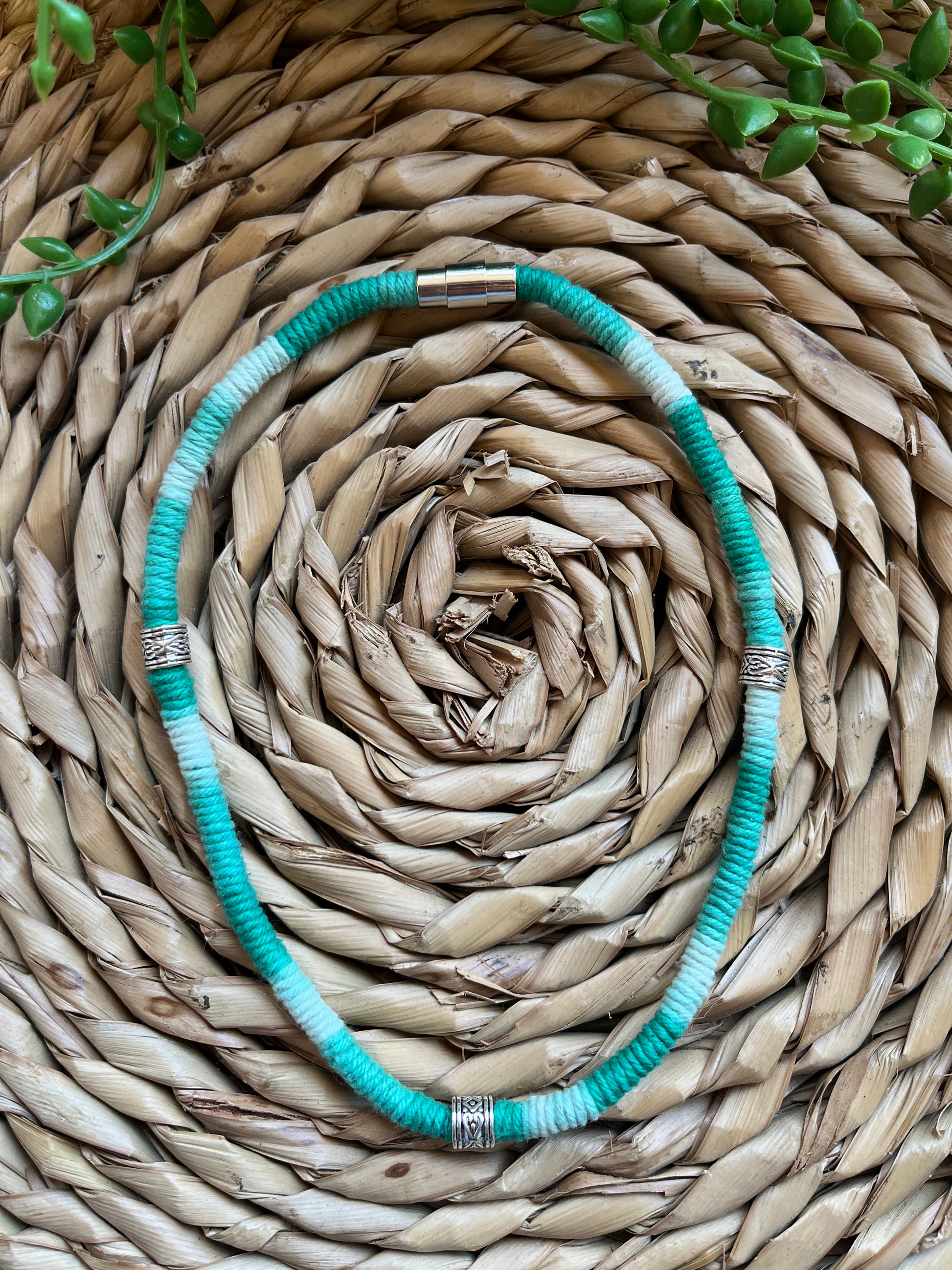 Handmade macrame surfer style short boho necklace, handmade using eco friendly green variegated yarn, and 3 Celtic style spacer beads. Displayed on a wicker mat next to some green plant vines. 