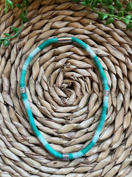 Handmade macrame surfer style short boho necklace, handmade using eco friendly green variegated yarn, and 3 Celtic style spacer beads. Displayed on a wicker mat next to some green plant vines. 