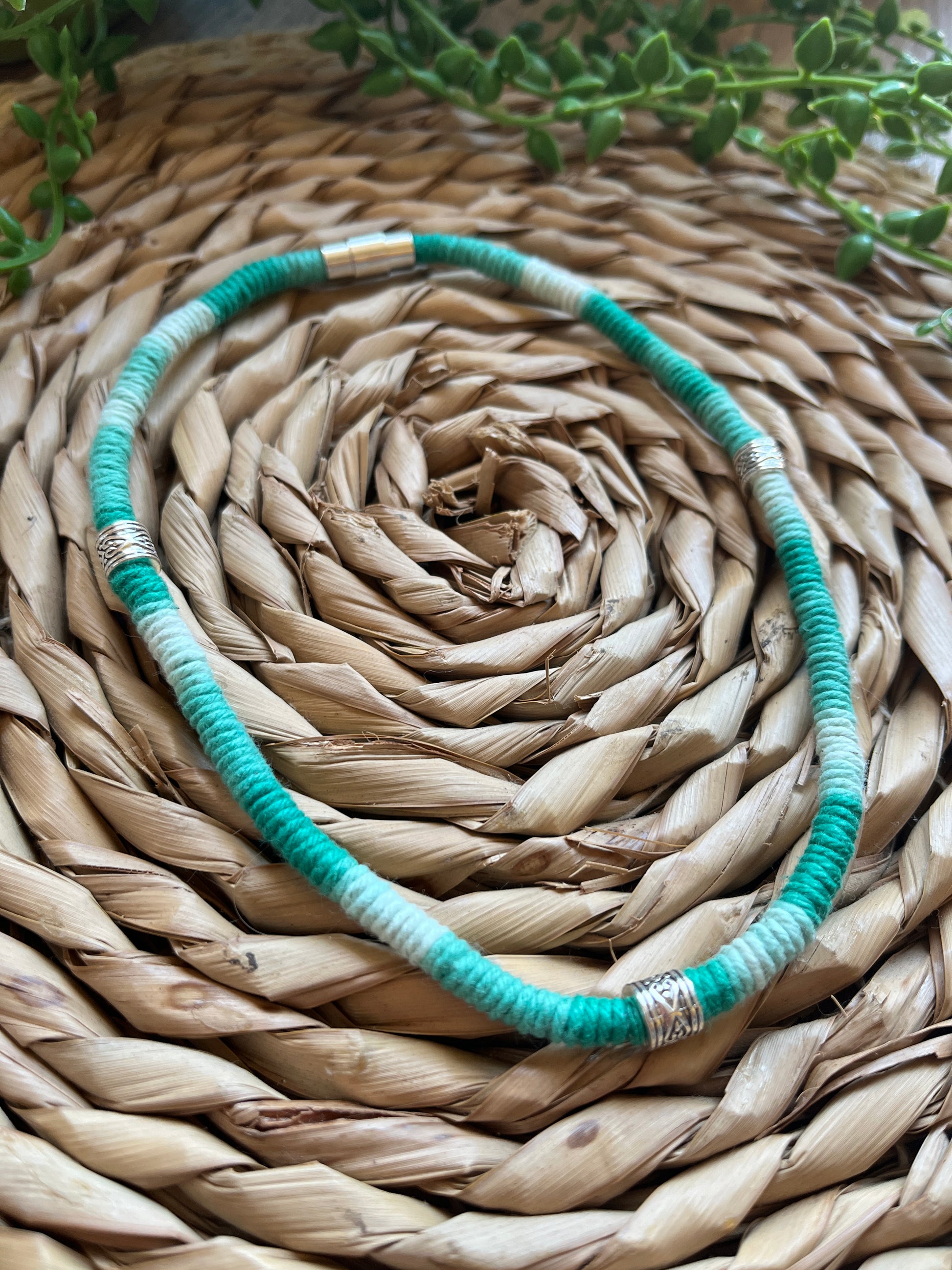 Handmade macrame surfer style short boho necklace, handmade using eco friendly green variegated yarn, and 3 Celtic style spacer beads. Displayed on a wicker mat next to some green plant vines. 