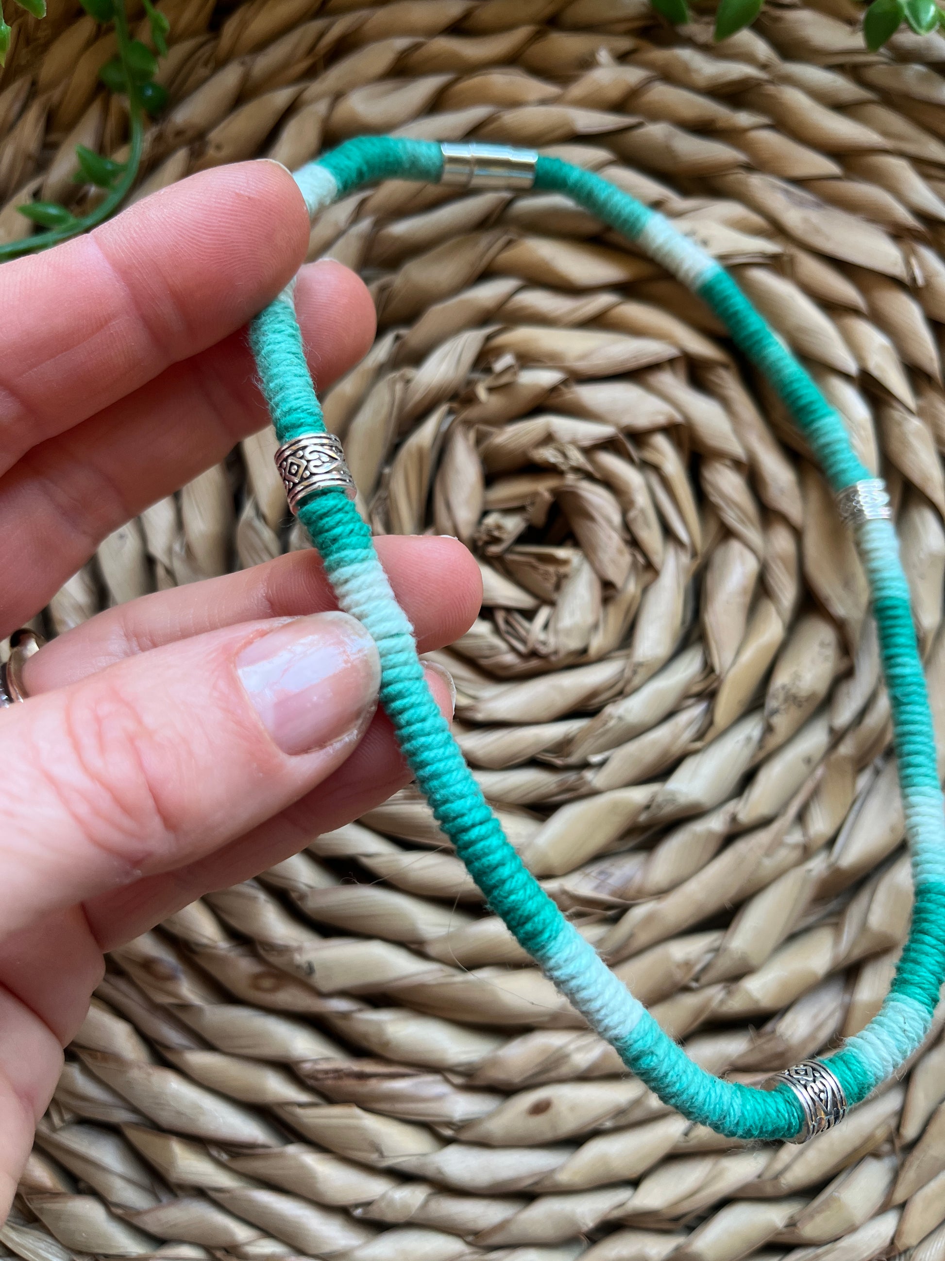 A hand holding a handmade macrame surfer style short boho necklace, handmade using eco friendly green variegated yarn, and 3 Celtic style spacer beads. Displayed on a wicker mat next to some green plant vines. 