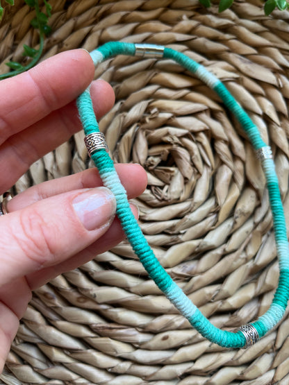 A hand holding a handmade macrame surfer style short boho necklace, handmade using eco friendly green variegated yarn, and 3 Celtic style spacer beads. Displayed on a wicker mat next to some green plant vines. 