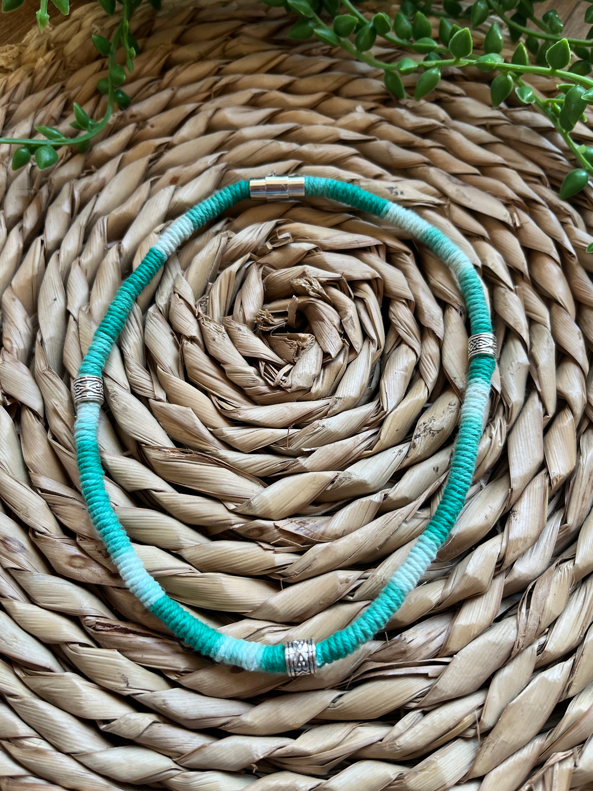 Handmade macrame surfer style short boho necklace, handmade using eco friendly green variegated yarn, and 3 Celtic style spacer beads. Displayed on a wicker mat next to some green plant vines. 
