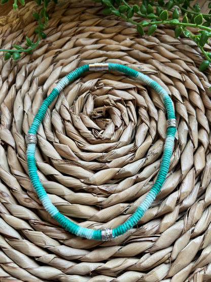 Handmade macrame surfer style short boho necklace, handmade using eco friendly green variegated yarn, and 3 Celtic style spacer beads. Displayed on a wicker mat next to some green plant vines. 