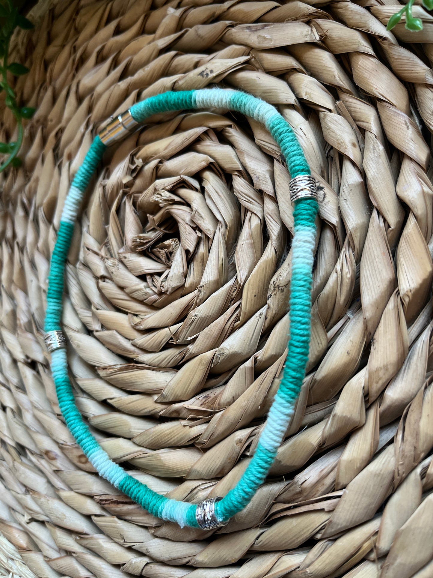 Handmade macrame surfer style short boho necklace, handmade using eco friendly green variegated yarn, and 3 Celtic style spacer beads. Displayed on a wicker mat next to some green plant vines. 