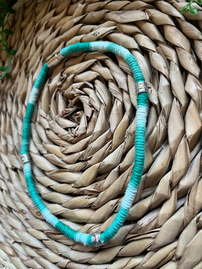 Handmade macrame surfer style short boho necklace, handmade using eco friendly green variegated yarn, and 3 Celtic style spacer beads. Displayed on a wicker mat next to some green plant vines. 