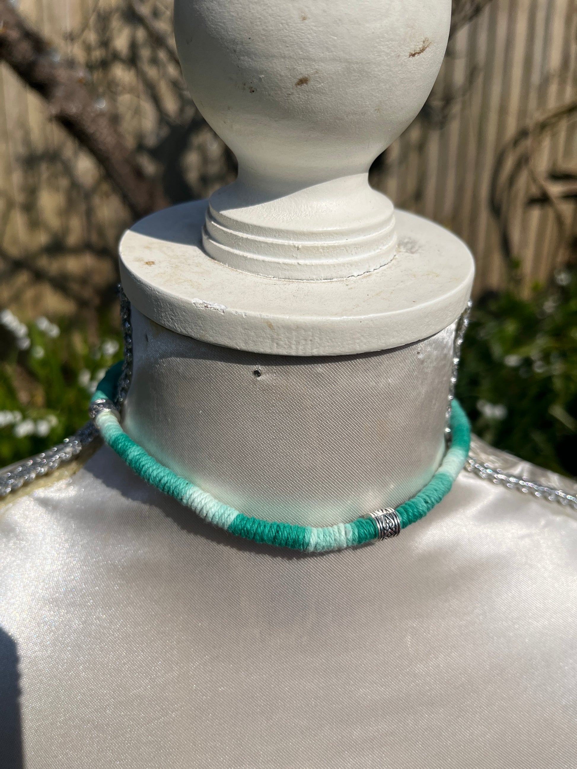 Handmade macrame surfer style short boho necklace, handmade using eco friendly green variegated yarn, and 3 Celtic style spacer beads. Displayed on a white mannequin, outside in the sunshine. 