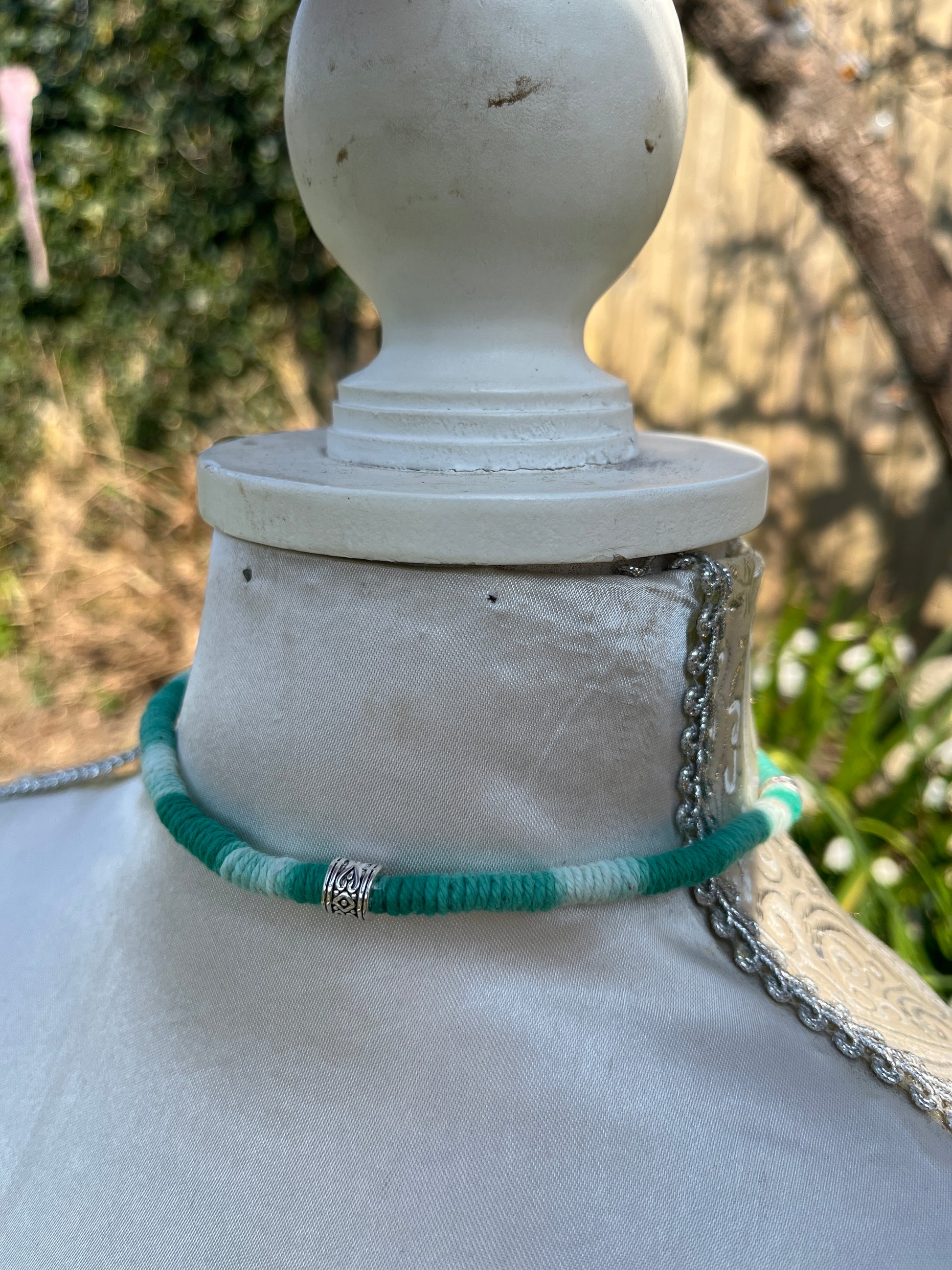 Handmade macrame surfer style short boho necklace, handmade using eco friendly green variegated yarn, and 3 Celtic style spacer beads. Displayed on a white mannequin, outside in the sunshine. 