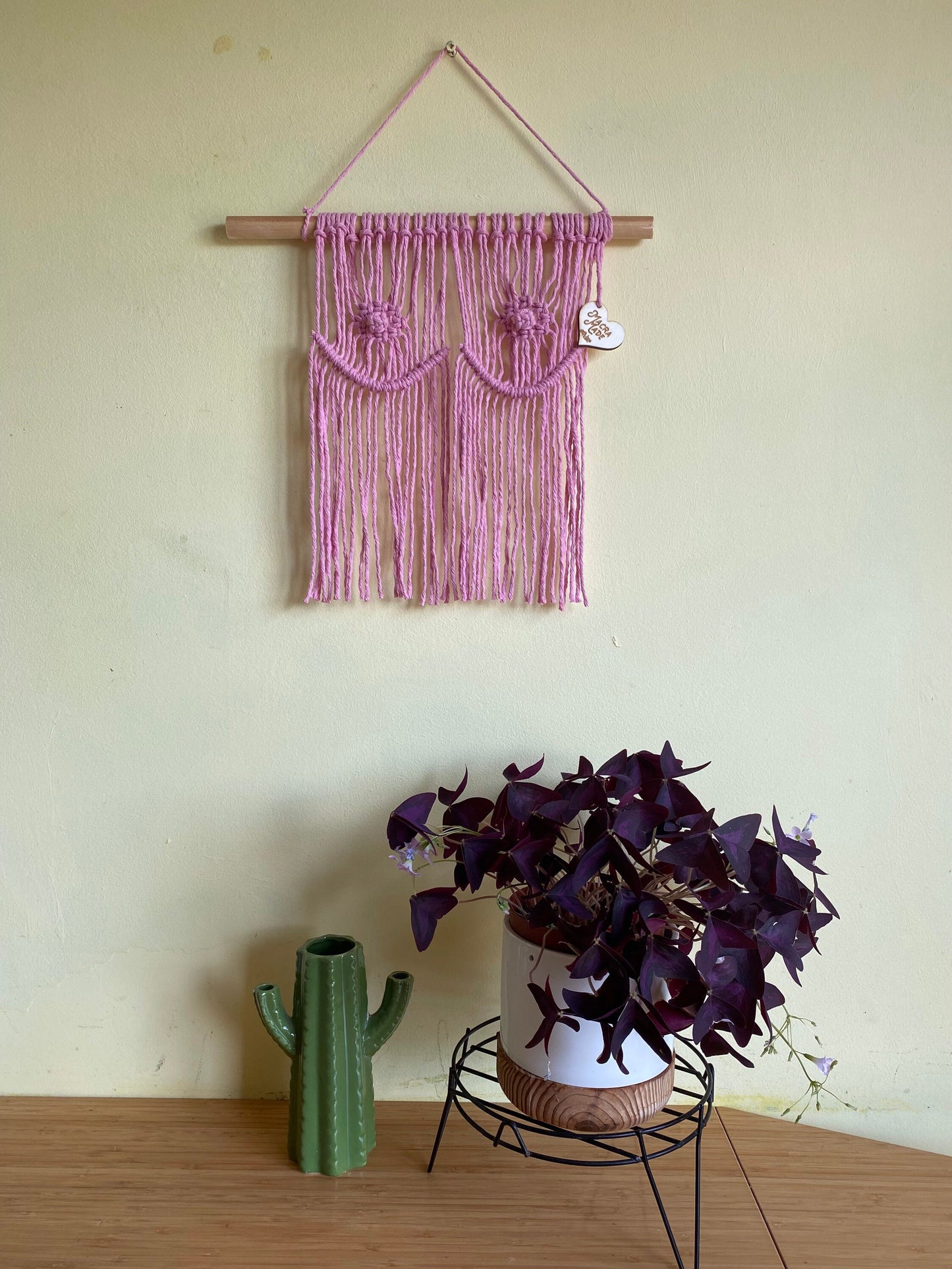 A pink, handmade Macra-Made-With-Love Unique colourful macrame boobs wall hanging decor 35x35cm with intricate patterns is displayed on a wooden dowel against a light yellow wall. Below, a white pot holding a plant with dark purple leaves and a cactus-shaped green ceramic vase sit on a wooden surface, adding to the boho-inspired decor.