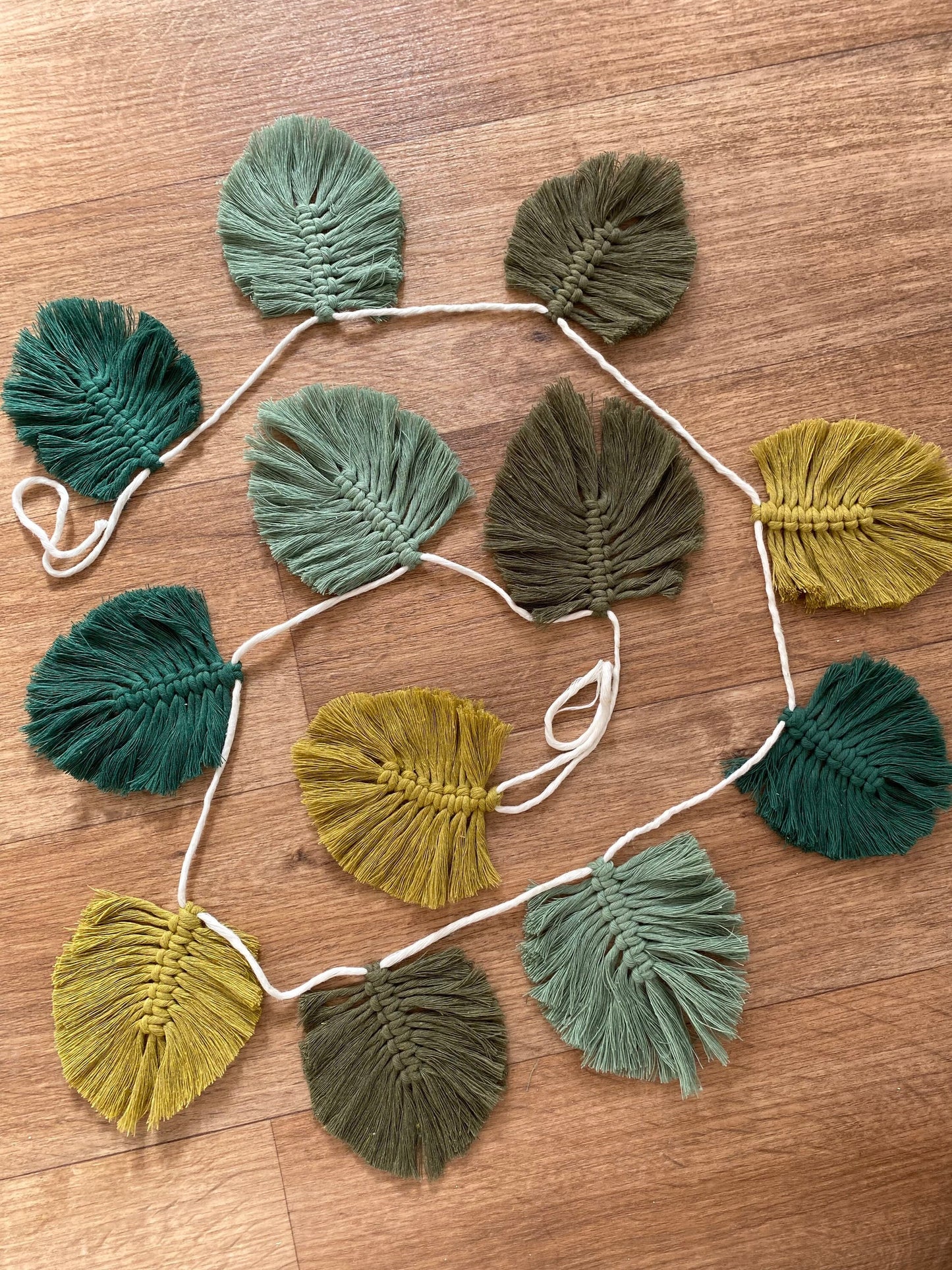 A handmade 100% cotton Autumn leaf garland - wall decor 180cm from Macra-Made-With-Love in various shades of green and yellow is laid out on a wooden floor. The leaves, attached to a white cord, create an autumnal wall hanging that showcases an assortment of textures and hues. This eco-friendly decor brings nature indoors beautifully.