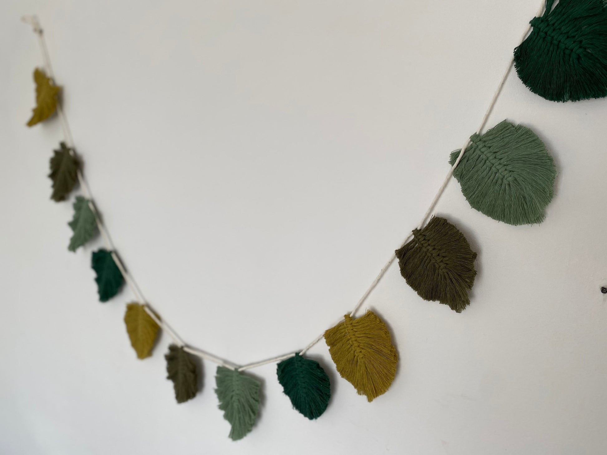 A handmade cotton garland of knitted leaves in various shades of green and yellow hangs on a white wall. This autumnal wall hanging, the 100% cotton Autumn leaf garland - wall decor 180cm by Macra-Made-With-Love, with its evenly spaced leaves along a thin string, creates a charming and eco-friendly decor that brings nature indoors.