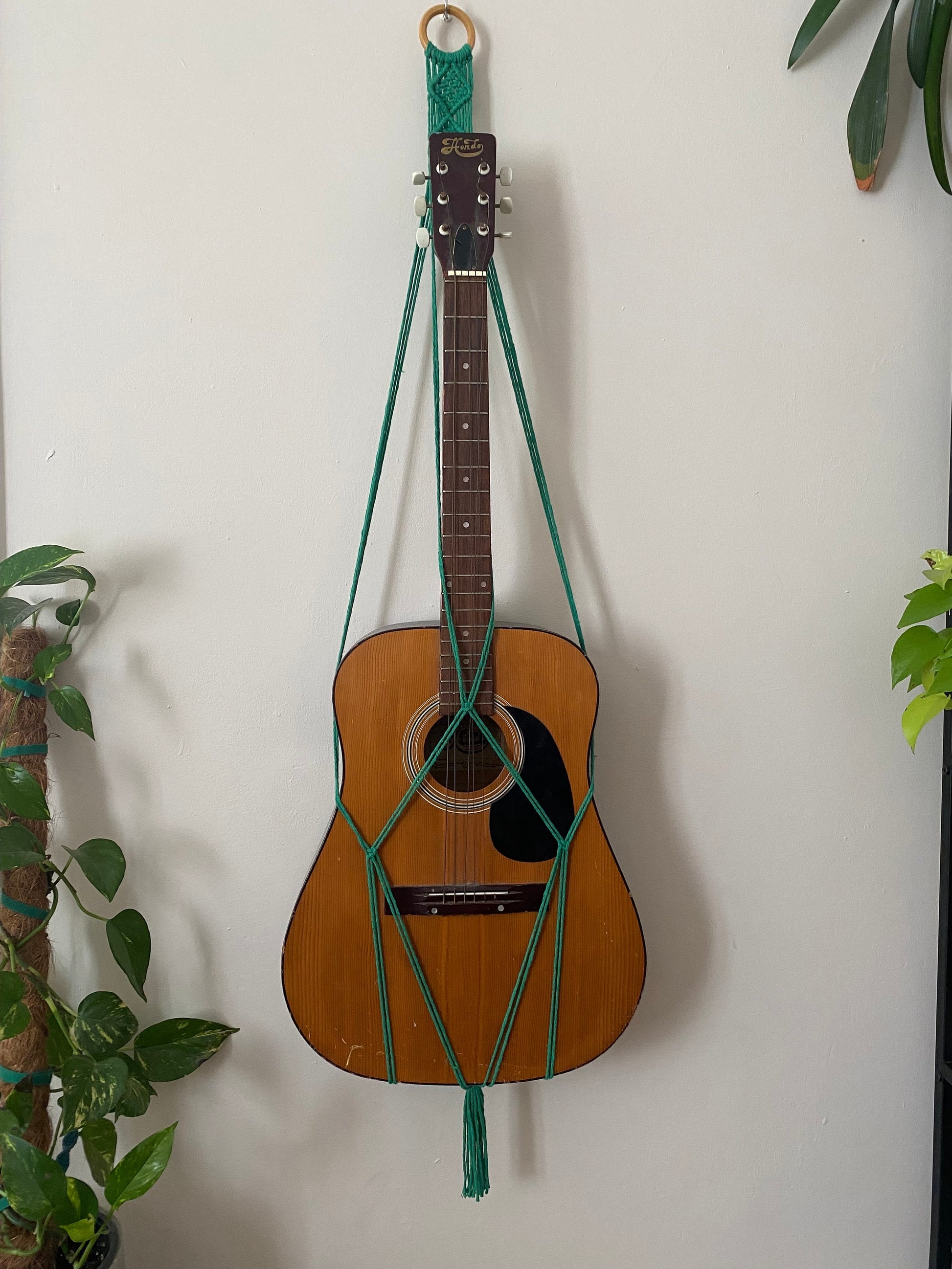 A brown acoustic guitar hangs on a white wall with green macrame cords, doubling as an eco-friendly guitar wall mount from Macra-Made-With-Love. The cords are intricately knotted, forming a triangular pattern. Green plants with lush leaves are partially visible on both sides of the image, adding to the boho chic home decor vibe.