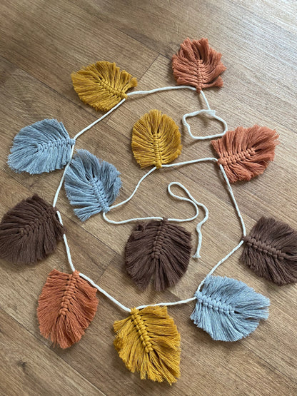 A Macra-Made-With-Love 100% cotton Autumn leaf garland - wall decor 180cm featuring macramé leaves of varying colors, including blue, yellow, brown, and orange, is laid out flat on a wooden surface. The leaves are attached to a white string, creating a colorful autumnal wall hanging that's perfect as eco-friendly decor.